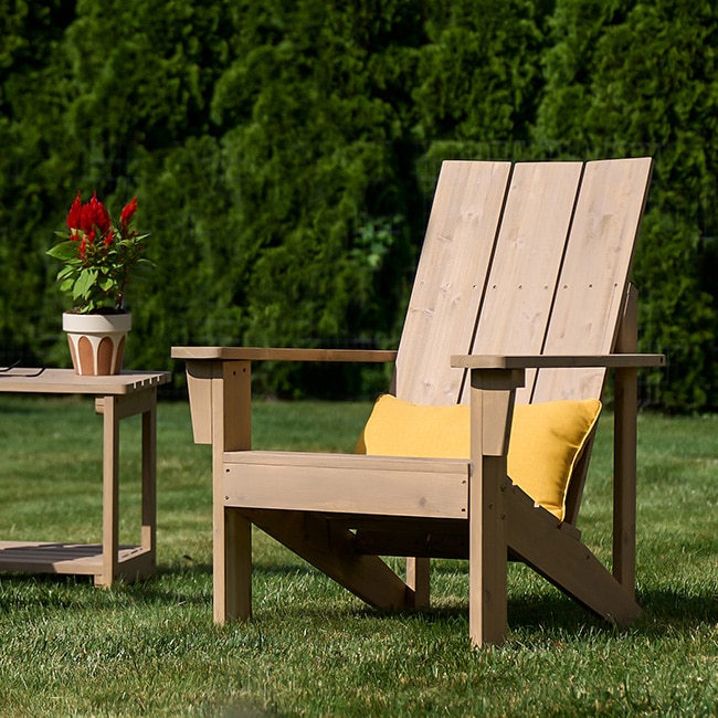 A lightly stained wooden Adirondack chair and a wood table sitting on a lush green lawn.