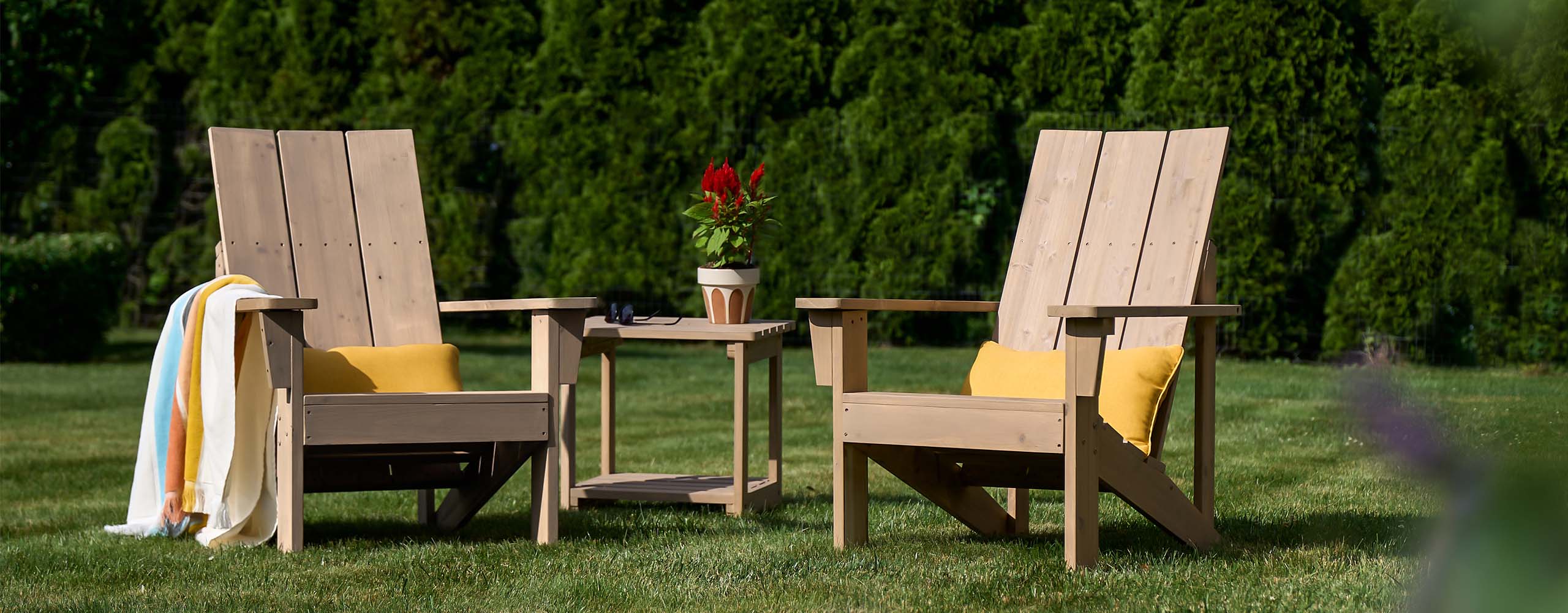 Two lightly stained wooden Adirondack chairs and a wood table sitting on a lush green lawn.