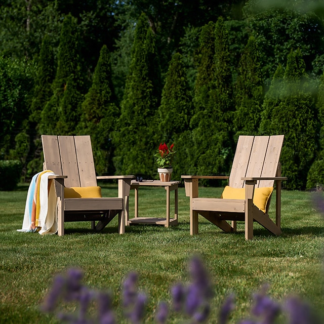 Two lightly stained wooden Adirondack chairs and a wood table sitting on a lush green lawn.