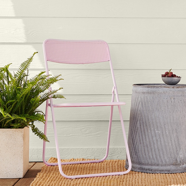 Two pink-painted metal folding chairs, a metal end table, and outdoor plant, in front of a white house with gray trim.