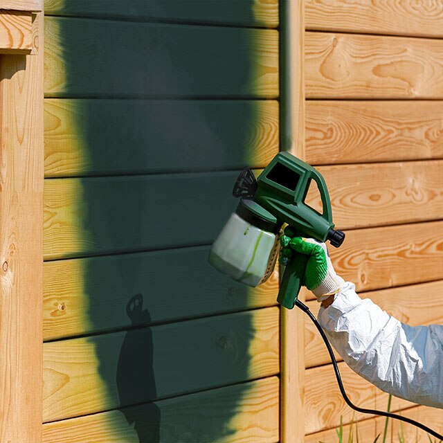 Un peintre recouvre de peinture verte la façade d’une maison au moyen d'un pulvérisateur.