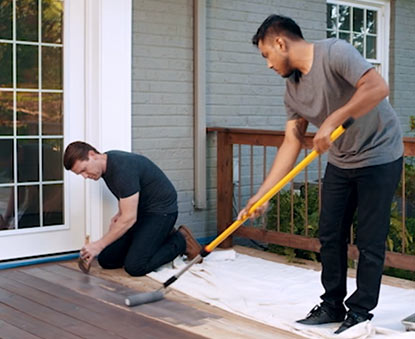 A gray brick home exterior with white trim and door has a deck being stained by two men: one man using a long roller, the other staining with a brush.