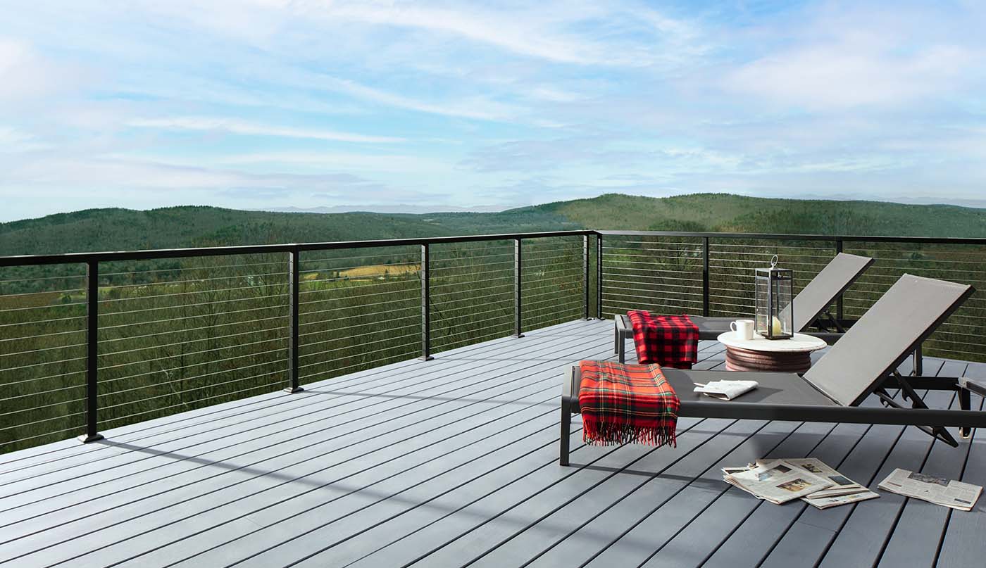 Two folding lounge chairs with draped red blankets on a light gray stained deck and a green mountainscape in the background.