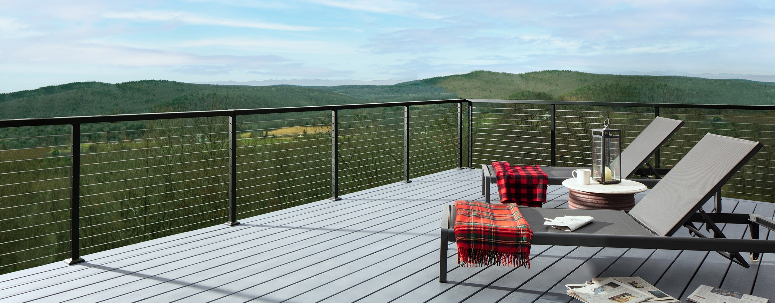 Two folding lounge chairs with draped red blankets on a light gray stained deck and a green mountainscape in the background.