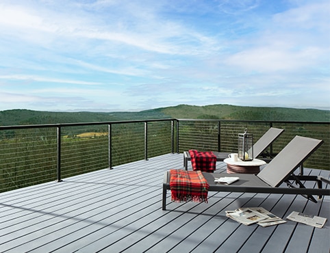 Two folding lounge chairs with draped red blankets on a light gray stained deck and a green mountainscape in the background.