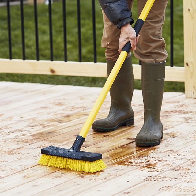 Un hombre con un largo cepillo para refregar amarillo que está preparando una nueva terraza de madera para teñirla y sellarla.