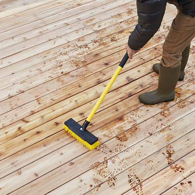 Un hombre con un largo cepillo para refregar amarillo que está preparando una nueva terraza de madera para teñirla y sellarla.