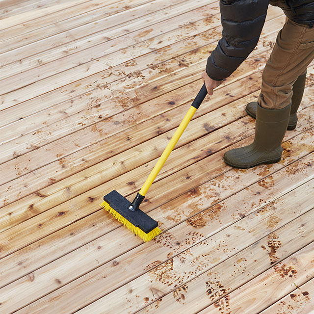 Un homme prépare la surface en bois d’une terrasse neuve avec une brosse jaune à long manche avant les travaux de scellement et de teinture.