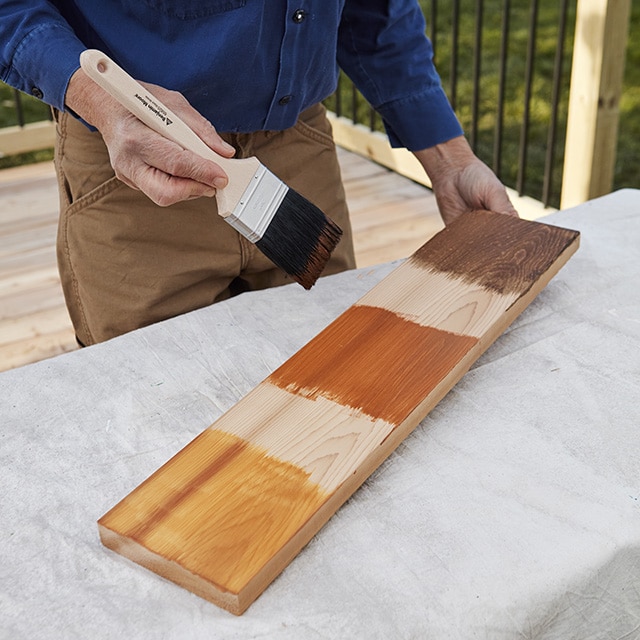 Un dueño de casa sosteniendo una brocha y una pieza de madera con tres muestras de tinte Woodluxe® aplicadas para probar el color.