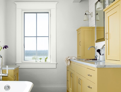 Une salle de bain moderne de style ferme avec des murs blanc cassé, des garnitures de fenêtre blanches et des armoires peintes en jaune doux avec des armoires en argent et des poignées de tiroir.