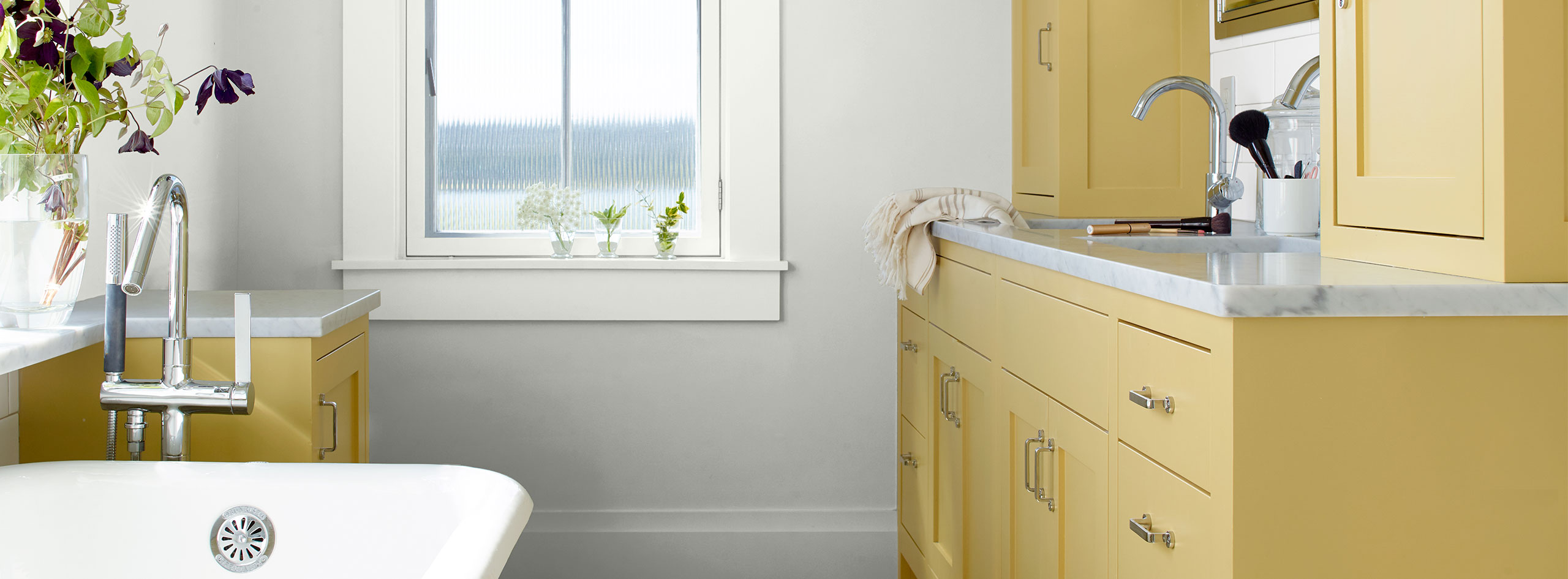 Une salle de bain moderne de style ferme avec des murs blanc cassé, des garnitures de fenêtre blanches et des armoires peintes en jaune doux avec des armoires en argent et des poignées de tiroir.
