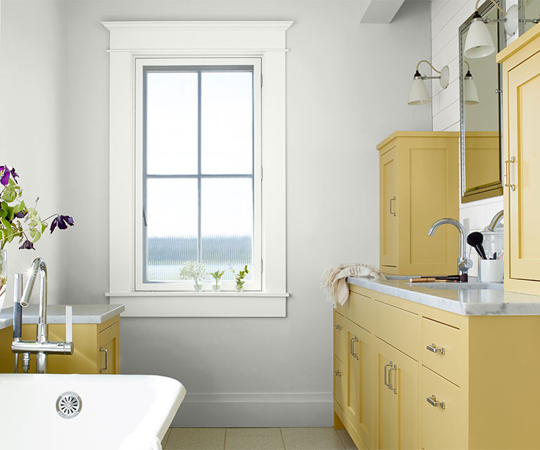 Une salle de bain moderne de style ferme avec des murs blanc cassé, des garnitures de fenêtre blanches et des armoires peintes en jaune doux avec des armoires en argent et des poignées de tiroir.