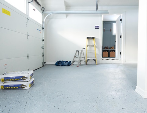 Un garage au plancher gris et aux murs blancs, un assortiment d’échelles, du ciment et une armoire.