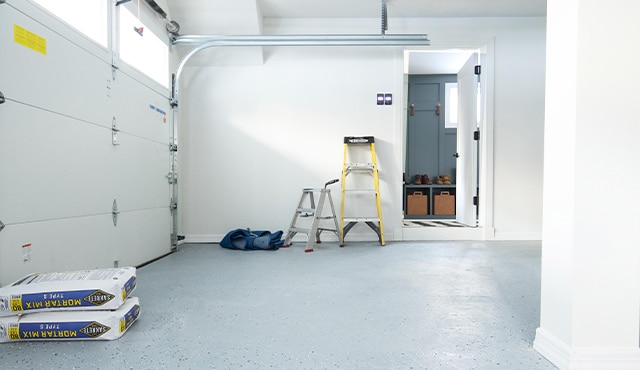 A garage with gray floor and white walls, an assortment of ladders, cement, and a cabinet.