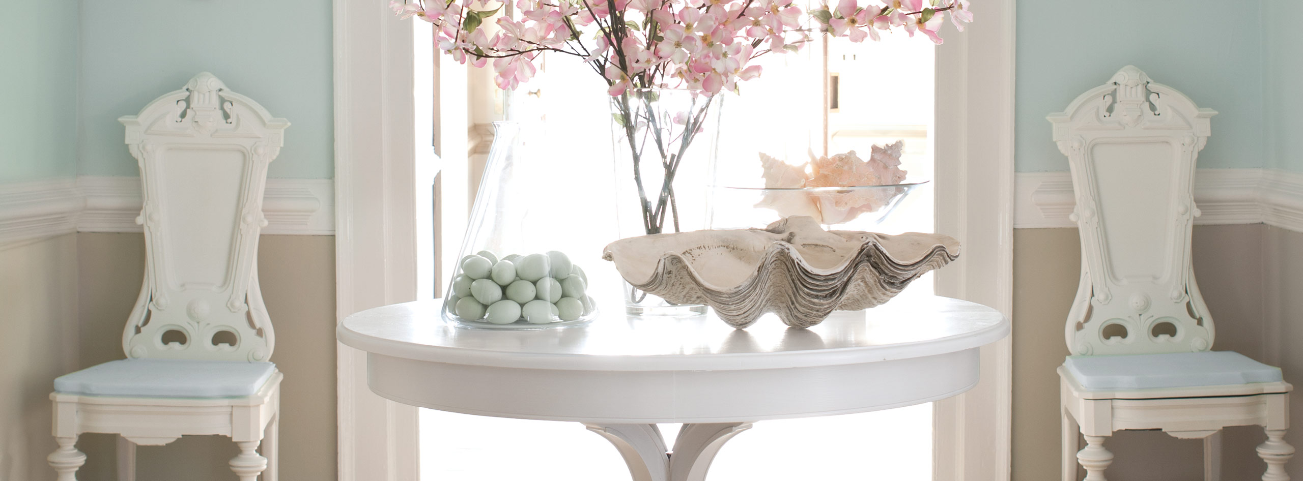 Un vestibule aux murs bleu clair ornés de moulures blanches et de lambris beiges, deux chaises blanches et une table peinturée en blanc sur laquelle sont posées des fleurs décoratives. 