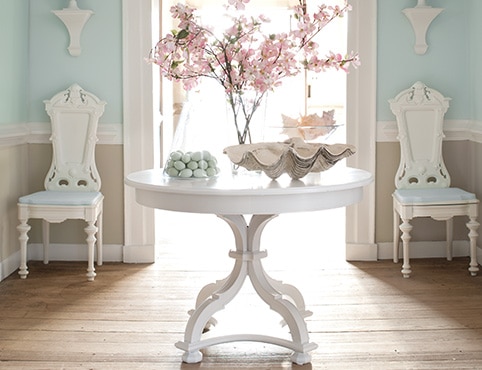 Un vestibule aux murs bleu clair ornés de moulures blanches et de lambris beiges, deux chaises blanches et une table peinturée en blanc sur laquelle sont posées des fleurs décoratives. 