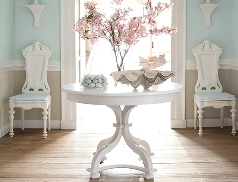 An entryway with a white-painted table with floral decor, two white chairs, and light blue walls, white trim, and beige wainscoting.