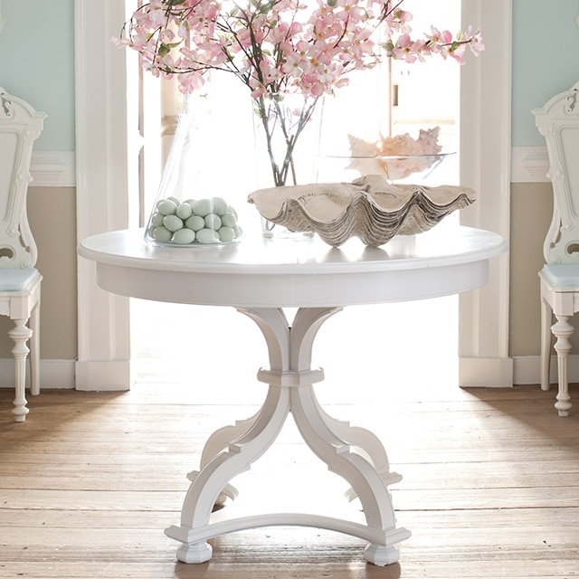 An entryway with a white-painted table with floral decor, two white chairs, and light blue walls, white trim, and beige wainscoting. 