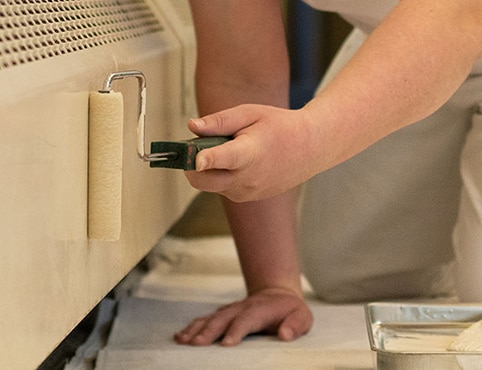 A person rolling off-white paint onto a baseboard heater.