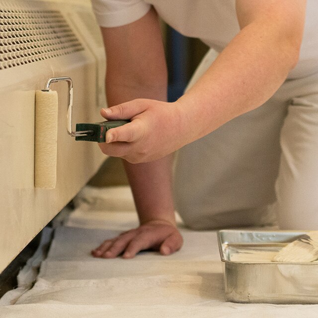 A person rolling off-white paint onto a baseboard heater.