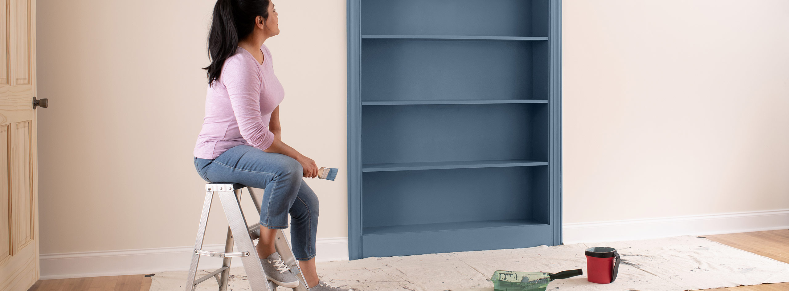 A homeowner sits on a step ladder in a pink-painted room, observing her freshly painted bookcase in a grayish blue.