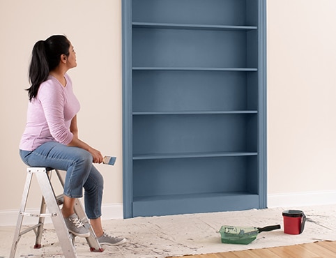 A homeowner sits on a step ladder in a pink-painted room, observing her freshly painted bookcase in a grayish blue.