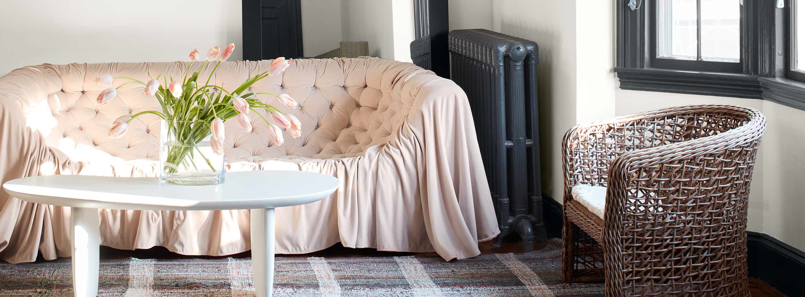 A white-painted living room with black trim and a black-painted radiator, a pink sofa, plaid rug, and modern chandelier.