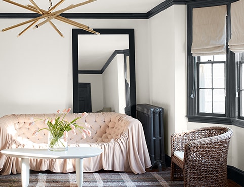 A white-painted living room with black trim and a black-painted radiator, a pink sofa, plaid rug, and modern chandelier.