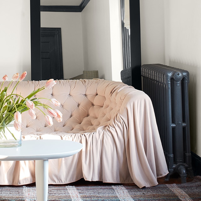 A white-painted living room with black trim and a black-painted radiator, a pink sofa, plaid rug, and modern chandelier.