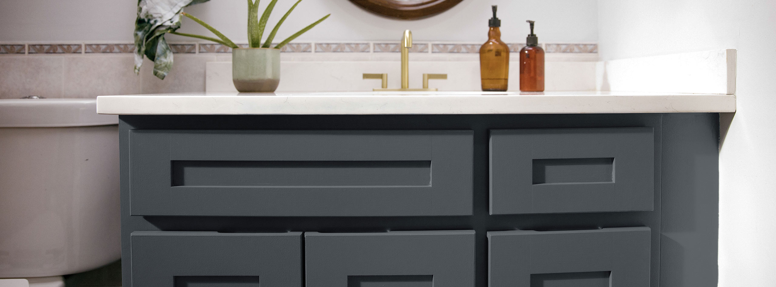 A close-up of a dark bathroom vanity cabinet, white countertops, muted pink tile backsplash, and one indoor plant.