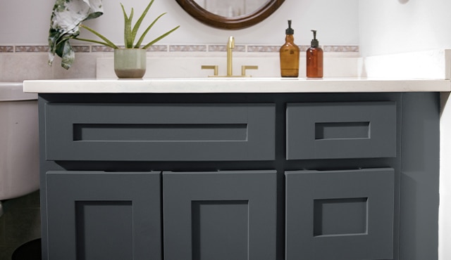 A close-up of a dark bathroom vanity cabinet, white countertops, muted pink tile backsplash, and one indoor plant.