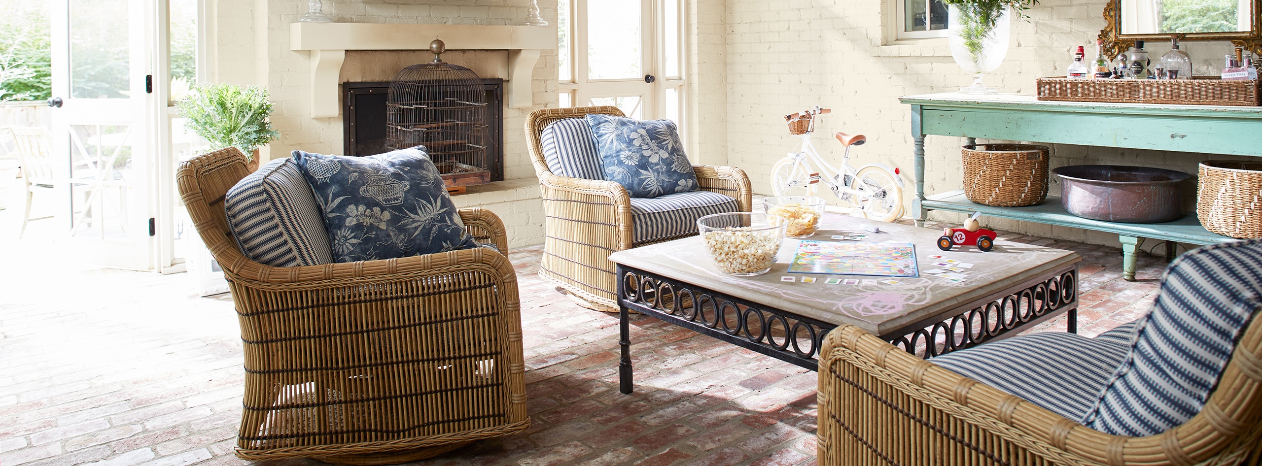 A boho-style living room with white-painted brick walls, wicker furniture, a wooden ceiling, and blue accent table. 