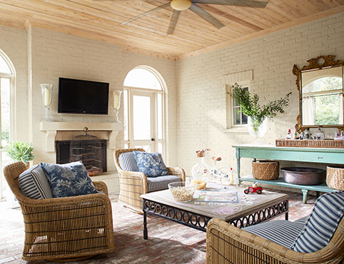 A boho-style living room with white-painted brick walls, wicker furniture, a wooden ceiling, and blue accent table. 