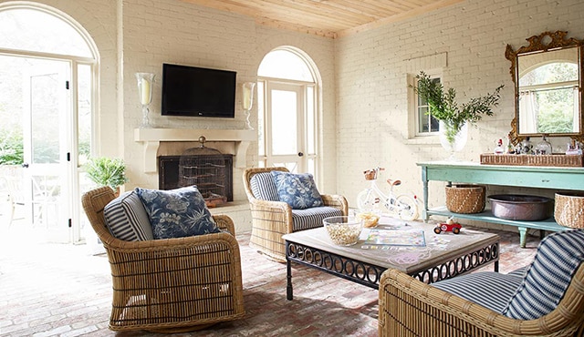 A boho-style living room with white-painted brick walls, wicker furniture, a wooden ceiling, and blue accent table. 