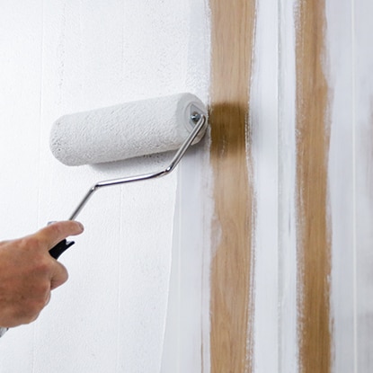 A homeowner rolls paint on a wood panelled wall. 