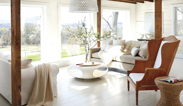 Un salon présentant des murs et un plancher peints en blanc, un fauteuil en bois, des canapés confortables et une table basse moderne.