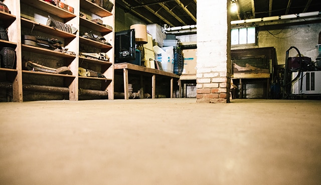 A concrete floor in a garage that needs to be sealed. A brick column is in the middle of the room, and wooden shelves line the walls.