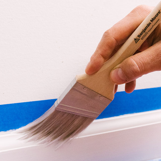 Guided by painter's tape, a homeowner uses a paintbrush to paint trim with white paint.