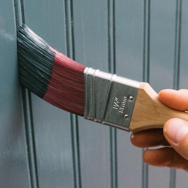 A homeowner using a paintbrush to paint a wall with grooves using a gray-blue paint.