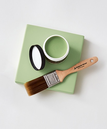 An open Benjamin Moore paint color sample and a paintbrush against a light green canvas.