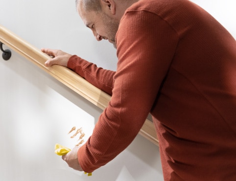 A person in a red shirt leaning against a handrail and holding a yellow cloth to clean a food stain off a white-painted staircase wall.