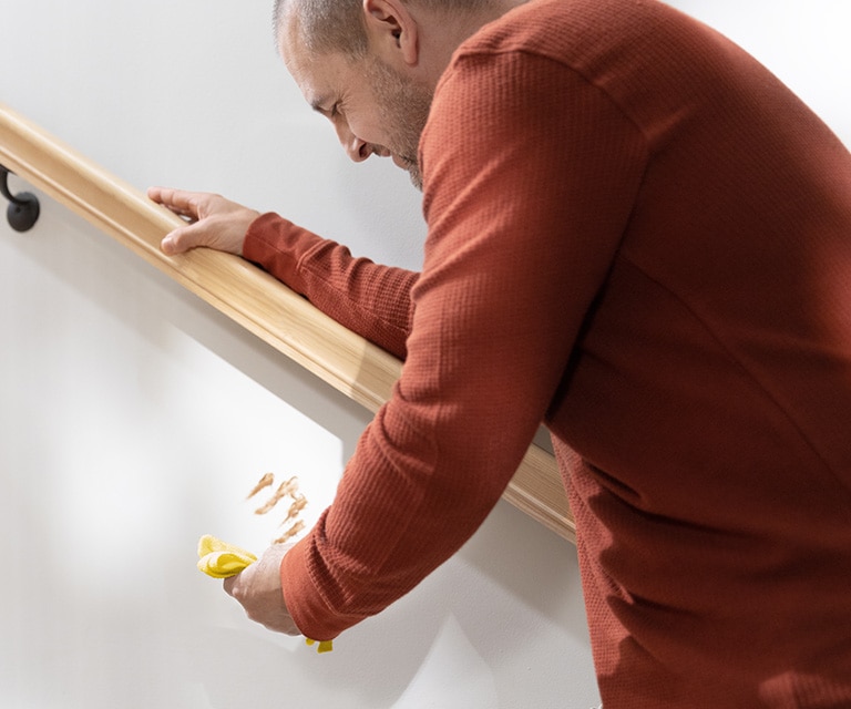 A person in a red shirt leaning against a handrail and holding a yellow cloth to clean a food stain off a white-painted staircase wall.