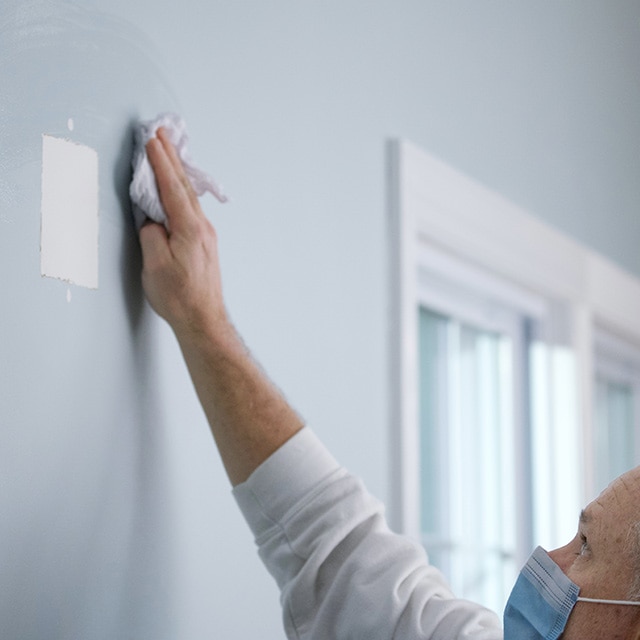 A homeowner wearing a mask wipes down a wall with a square patch.