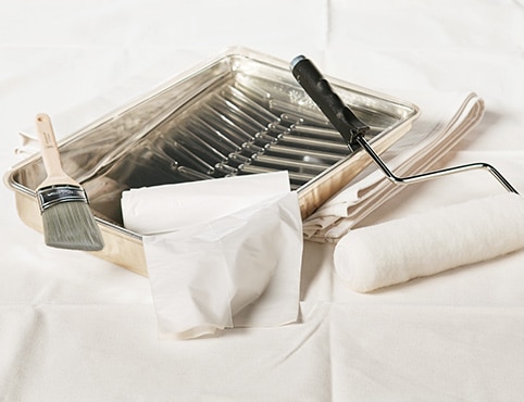 Painting supplies including a metal paint tray, an angled sash brush, a paint roller and a plastic drop cloth.