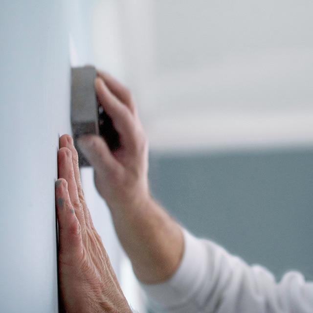 A homeowner wearing a mask sands down a wall.