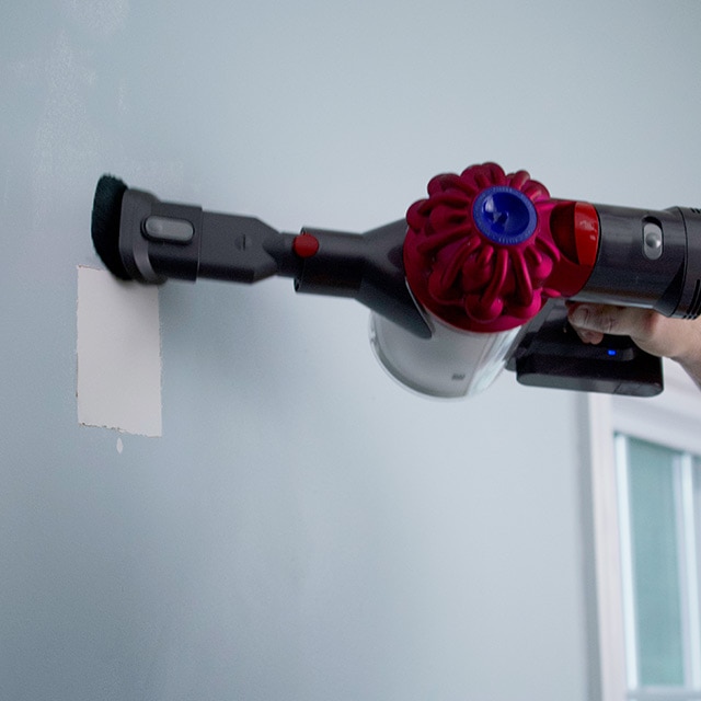 A homeowner wearing a mask vacuums dust off of a wall with a square patch.