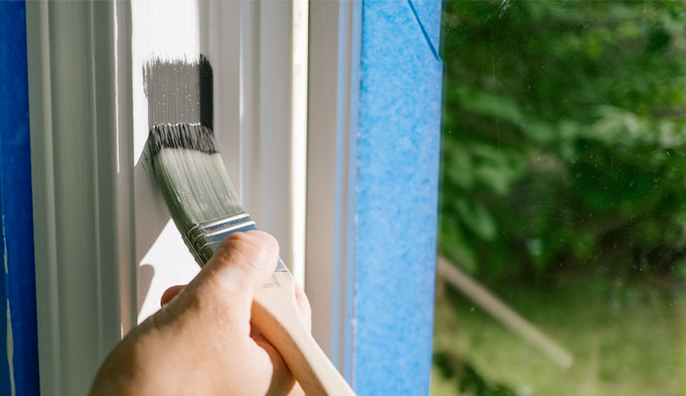 A homeowner painting white window trim with gray paint.