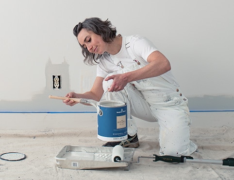 A woman holding a 3.79 L can of paint preparing to paint a wall.