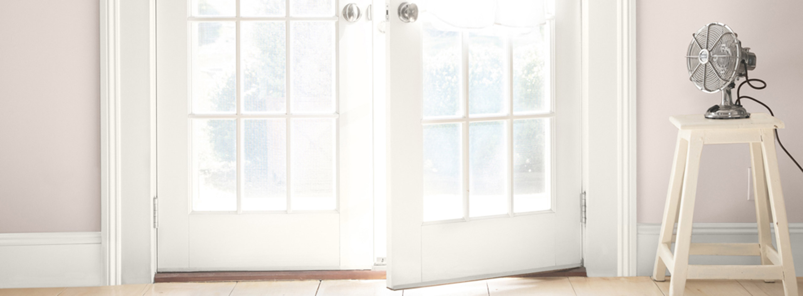 White-painted French doors opening in a pinkish-beige room. A fan sits on a white stool, and a white button-up is hanging on a door.