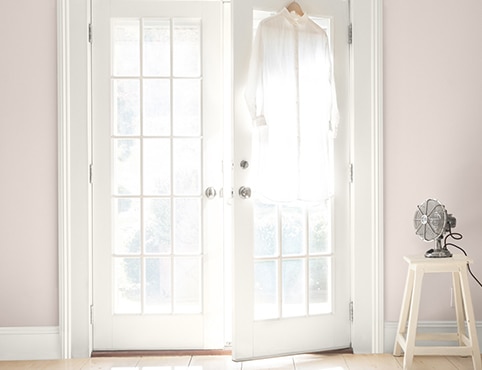 White-painted French doors opening in a pinkish beige room. A fan sits on a white stool, and a white button up is hanging on a door.